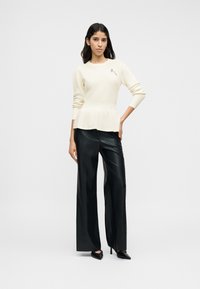 Κρεμ πλεκτό πουλόβερ με πεπλούμ και μακριά μανίκια, διακοσμημένο με ένα διακοσμητικό κονκάρδα. Συνδυασμένο με μαύρο δερμάτινο παντελόνι υψηλής μέσης και φαρδιάς γραμμής.