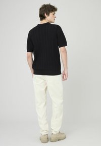 Polo noir en maille avec texture côtelée et manches courtes, associé à un pantalon crème et des chaussures beiges. Vue arrière mise en avant.