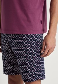 Lila kurzärmliges Hemd kombiniert mit marineblauen gemusterten Shorts, die Rauten in Pink und Weiß zeigen. Glatte Textur, lässiges Design.