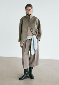 Brown wool coat with a high collar and black buttons, beige sweater, light blue knitted wrap, beige wide-leg pants, and black ankle boots.