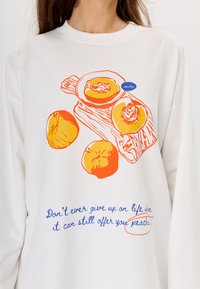 Witte sweatshirts met oranje en gele perzikgraphics en blauwe tekst. De tekst luidt: "Geef nooit op het leven wanneer het je nog perziken kan bieden."