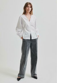Camicia bianca con scollo a V profondo, maniche a palloncino e tessuto strutturato. Abbinata a jeans grigi a vita alta con gamba larga e tacchi neri.