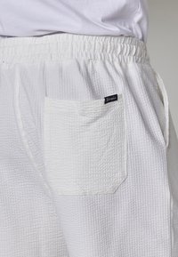 Primo piano di pantaloni bianchi con texture, dotati di una cintura elastica e tasca posteriore quadrata con un piccolo etichetta del marchio nera.