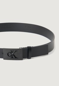 Curea din piele neagră, cu o cataramă rectangulară mată și un logo „cK” embosat. Textură netedă și design al curelei ajustabil.