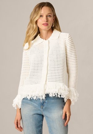 Blonde woman wearing hoop earrings, white knit cardigan with open weave and fringed hem, paired with light blue jeans.