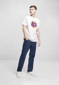 Hvit T-skjorte med korte ermer og "TUNE SQUAD" grafikk i blått og oransje, parret med mørkeblå denimjeans og hvite sneakers.
