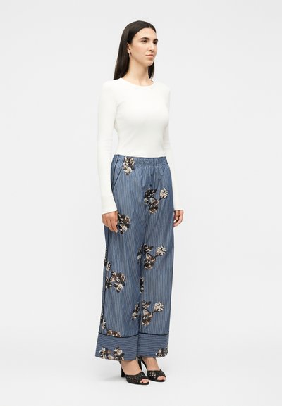 Femme portant un haut blanc à manches longues, un pantalon large bleu avec des motifs floraux et à rayures, et des talons ouverts noirs, debout de côté.