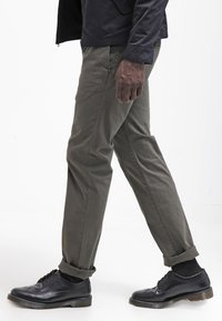 Pantaloni di cotone verde oliva con risvolti, indossati con scarpe brogue in pelle nera lucida con perforazioni decorative.