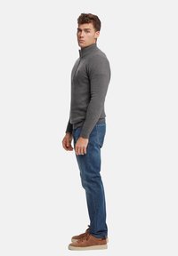 Suéter gris con cremallera y puños acanalados, combinado con jeans azules y zapatillas de gamuza marrón, visto de lado. Detalles de tela texturizada visibles.