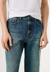 Blå denim jeans med en falmet tekstur, fem-lomme design og brune syninger. Sammen med en hvid kortærmet skjorte.