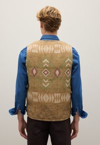 Wollweste in Tan mit geometrischen Mustern in Beige, Grün und maroonfarben, getragen über einem blauen Denimhemd. Glatte Textur und taillierter Schnitt.