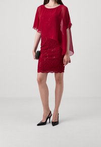 Czerwona sukienka z przezroczystą nakładką ozdobioną cekinami. Czarne czółenka na pasku z tyłu i mała czarna kopertówka. Gładka tekstura i dopasowany krój.