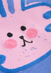 Roza tkaninski zajčkov obraz z črnimi očmi, nosom in usti, rožnatimi ličnicami ter modrim vezavilom na modrem tekstilnem ozadju.