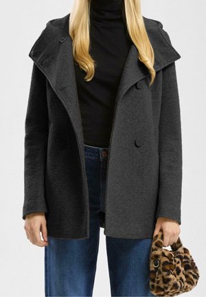 Vrouw draagt een donkergrijze getextureerde dubbelgeknoopte jas, zwarte coltrui, blauwe spijkerbroek, houdt een kleine handtas van nepbont met luipaardprint vast.