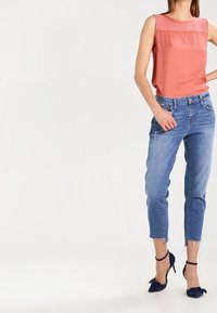 Ljus korallfärgad utan ärmar-top med spetsdetaljer, matchad med ljusblå slitna jeans och marinblå klackskor med rosettaccenter.