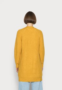 Cardigan giallo lungo con lavorazione a maglia testurizzata, collo rotondo e spalle scese, che si estende oltre i fianchi, abbinato a jeans azzurri chiari.