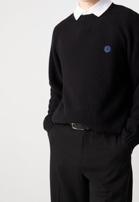 Pull noir tricoté avec manches raglan, poignets côtelés et encolure ronde. Présente un petit logo smiley bleu sur la poitrine. Associé à un pantalon noir.