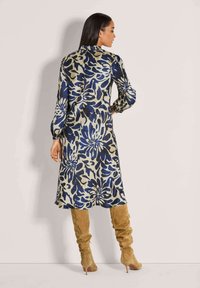 Frau mit langen Haaren, die ein langärmeliges, knielanges Blumenkleid und beige kniehohe Stiefel mit Absatz trägt, schaut gegen eine Wand.