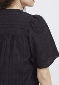 Blusa nera ricamata con un motivo floreale testurizzato, maniche puff e una spalla con dettagli di cucitura orizzontale.