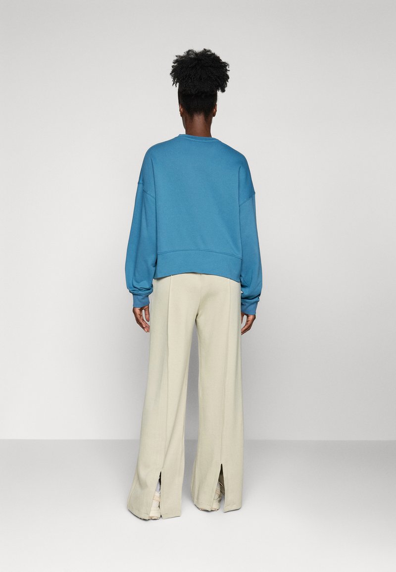 Felpa blu oversize con spalle cadute e orlo a coste, abbinata a pantaloni beige a gamba larga con spacchi alla caviglia.