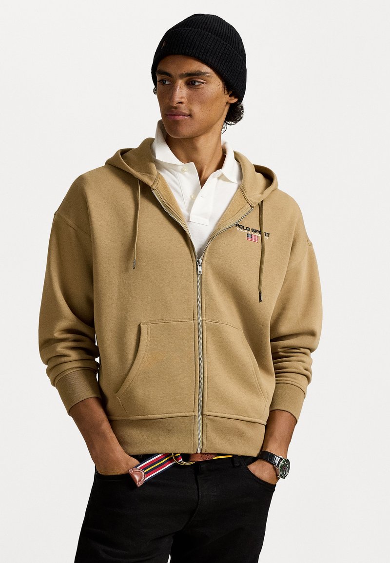 Sweatshirt com fecho éclair castanho feito de tecido macio, com um bolso frontal e capuz com cordão. Usado por cima de uma camisa polo branca. Gorro preto.