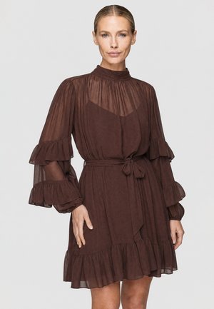 Frau trägt ein braunes transparentes Kleid mit langen Ärmeln und Rüschen an Ärmeln und Saum, mit gebundener Taille, steht vor schlichtem Hintergrund.