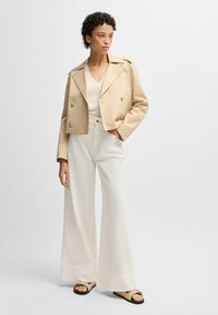 Beige kort dubbelknäppt jacka med två knappar, över en vit topp. Matchad med vida vita byxor och beiga sandaler.