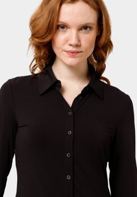 Černá košile na knoflíky s dlouhými rukávy a límcem. Hladká látka s vypasovaným střihem a kontrastními knoflíky vpředu.