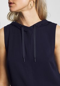 Haut sans manches bleu marine avec capuche, présentant un cordon de serrage avec des embouts cylindriques, une texture de tissu lisse et un design minimaliste.