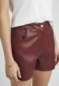 Shorts en simili cuir bordeaux avec une texture lisse, poches avant et fermeture à bouton argenté. Coupe ajustée avec un design épuré.