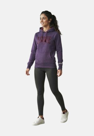 Donna con capelli scuri raccolti in una coda di cavallo, indossa una felpa con cappuccio viola, leggings grigio scuro e scarpe da ginnastica bianche, in piedi con un piede leggermente sollevato.