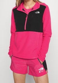 Fleece-pullover i starkrosa med svarta detaljer, design med kvart dragkedja och logotyp. Matchas med rosa shorts med fickor.