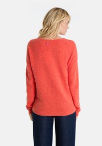 Korallfarbener Wollpullover mit lässiger Passform, langen Ärmeln und einem Rückenausschnitt, der durch einen kleinen rosa Knopf betont wird. Kombiniert mit dunklen Jeans.
