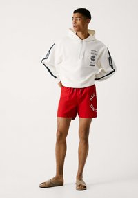 Sudadera blanca con estampados de logotipos, combinada con bañadores rojos que presentan el texto "RED". El modelo lleva sandalias beige. Look simple y deportivo.