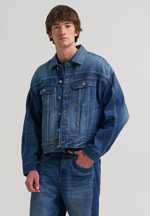 Jeune homme portant une veste en jean bleue et un jean, debout avec une main dans la poche sur un fond clair uni.