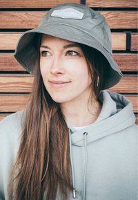 Olivgrön bucket hat med en bred brätte, med en texturerad tyg och en rektangulär patch på framsidan. Bärs med en ljusgrå hoodie.