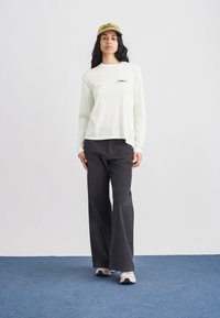 Lichtgroen shirt met lange mouwen en 'OBEY' opdruk; donkergrijze broek met wijde pijpen; gele pet met afbeelding; witte en grijze sneakers; effen achtergrond.