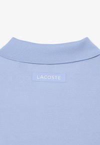 Tela texturizada azul claro con un cuello acanalado y una etiqueta de la marca Lacoste centrada debajo del cuello.