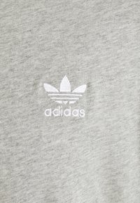Ljust grå bomullströja med en vit broderad Adidas-logga och en trefoil-design ovanför varumärket. Mjuk textur.