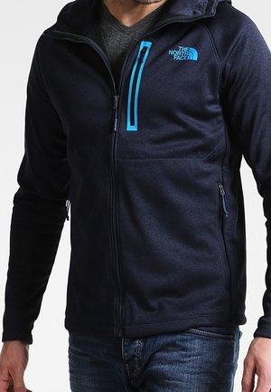 Man draagt een marineblauwe The North Face jas met rits en blauwe ritsdetails over een grijs shirt en een blauwe spijkerbroek.