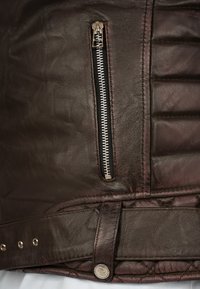 Giacca in pelle marrone con una tasca con zip argento, texture trapuntata e dettaglio con bottone a pressione in vita con accenti metallici.