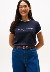 Granatowy bawełniany t-shirt z białymi lamówkami, z logo "TOMMY HILFIGER EST. 1985". Połączony z dżinsami i brązowym paskiem.