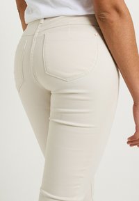Jeans skinny beige con una textura suave, que cuentan con dos bolsillos en la parte posterior y una cintura alta. El diseño mantiene una silueta ajustada y elegante.