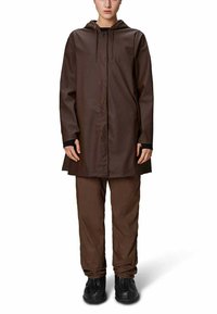 Cappotto impermeabile marrone con cappuccio, chiusura frontale a pressione e tasche laterali, abbinato a pantaloni marroni coordinati e scarpe nere.