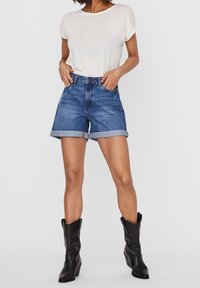 Denimshorts mit umgeschlagenen Säumen, blauer Farbe, hochgeschnittenes Design, kombiniert mit einem weißen T-Shirt und schwarzen Cowboy-Stiefeln. Glatte Textur.