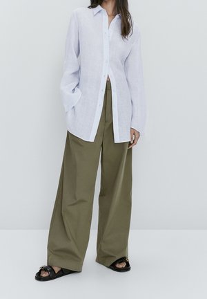 Femme portant une chemise blanche ample boutonnée, un pantalon large vert olive et des sandales noires, debout devant un fond clair uni.