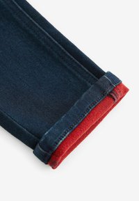 Un jean en denim bleu foncé avec un revers rouge. Le tissu est robuste avec des coutures visibles le long du revers. L'ourlet roulé révèle une couleur contrastante.
