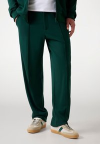 Donkergroene joggingsbroek met een gladde, rekbare stof en een slank ontwerp, met geplooide voorkant en zijzakken, gecombineerd met gebroken witte sneakers.