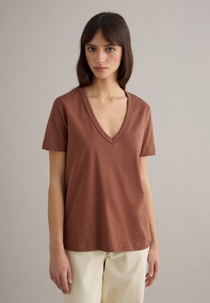 Donna con capelli castani lisci, che indossa una maglietta marrone a maniche corte con scollo a V e pantaloni color crema, in piedi davanti a uno sfondo semplice grigio chiaro.