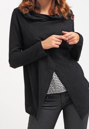 Femme portant un pull noir à manches longues avec des boutons, tenant le devant ouvert pour montrer une chemise à motif noir et blanc en dessous.
